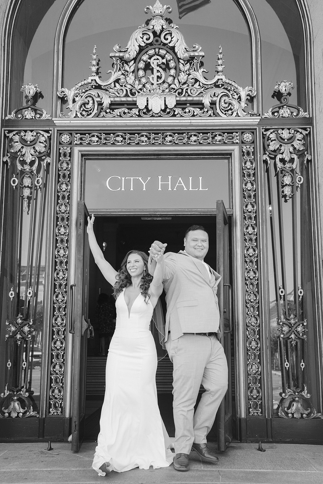 Black-and-white photograph of a bride and groom celebrating together in front of the doors at San Francisco City Hall.