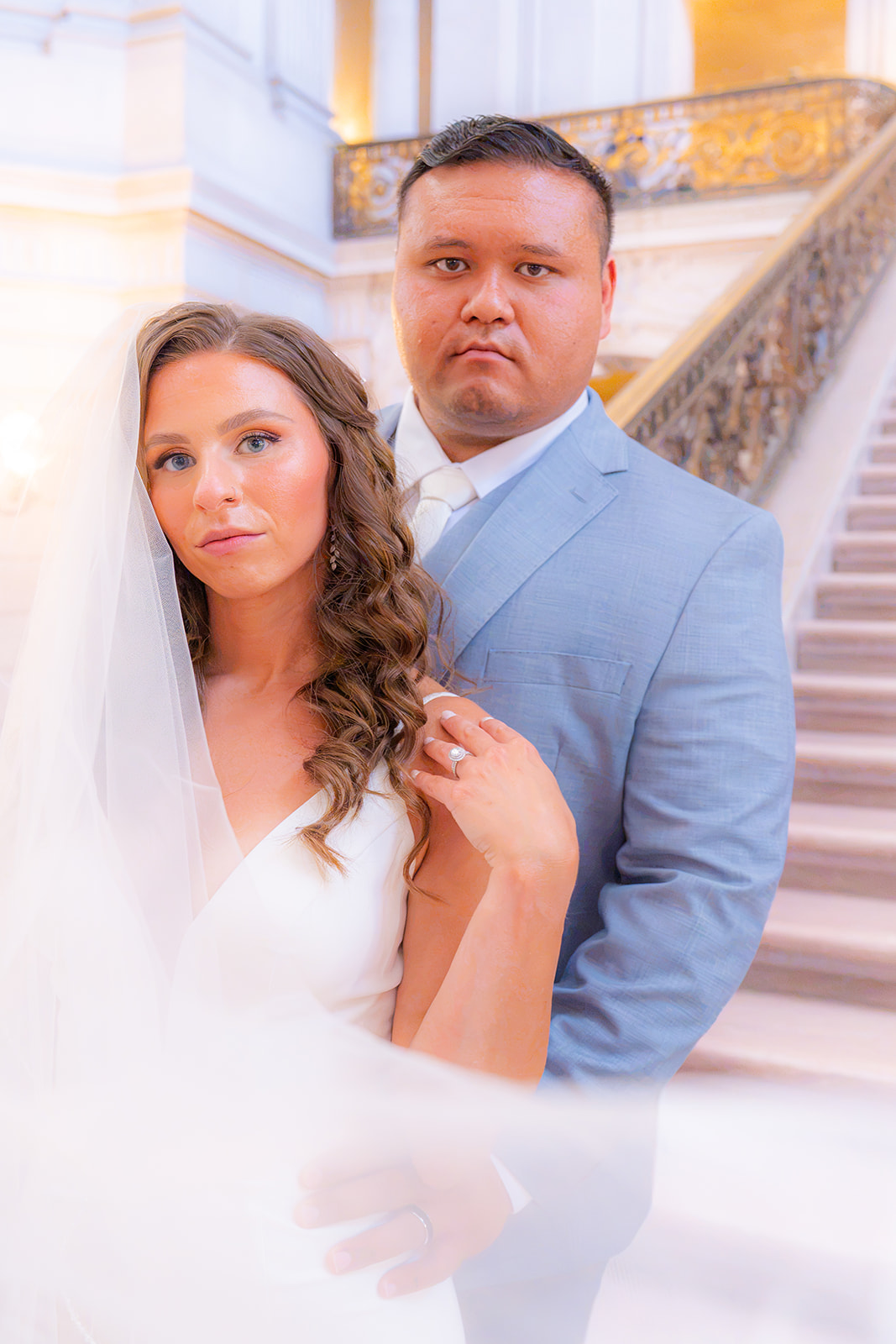 Bride and groom posing together with the bride’s wedding veil on a staircase at San Francisco City Hall.