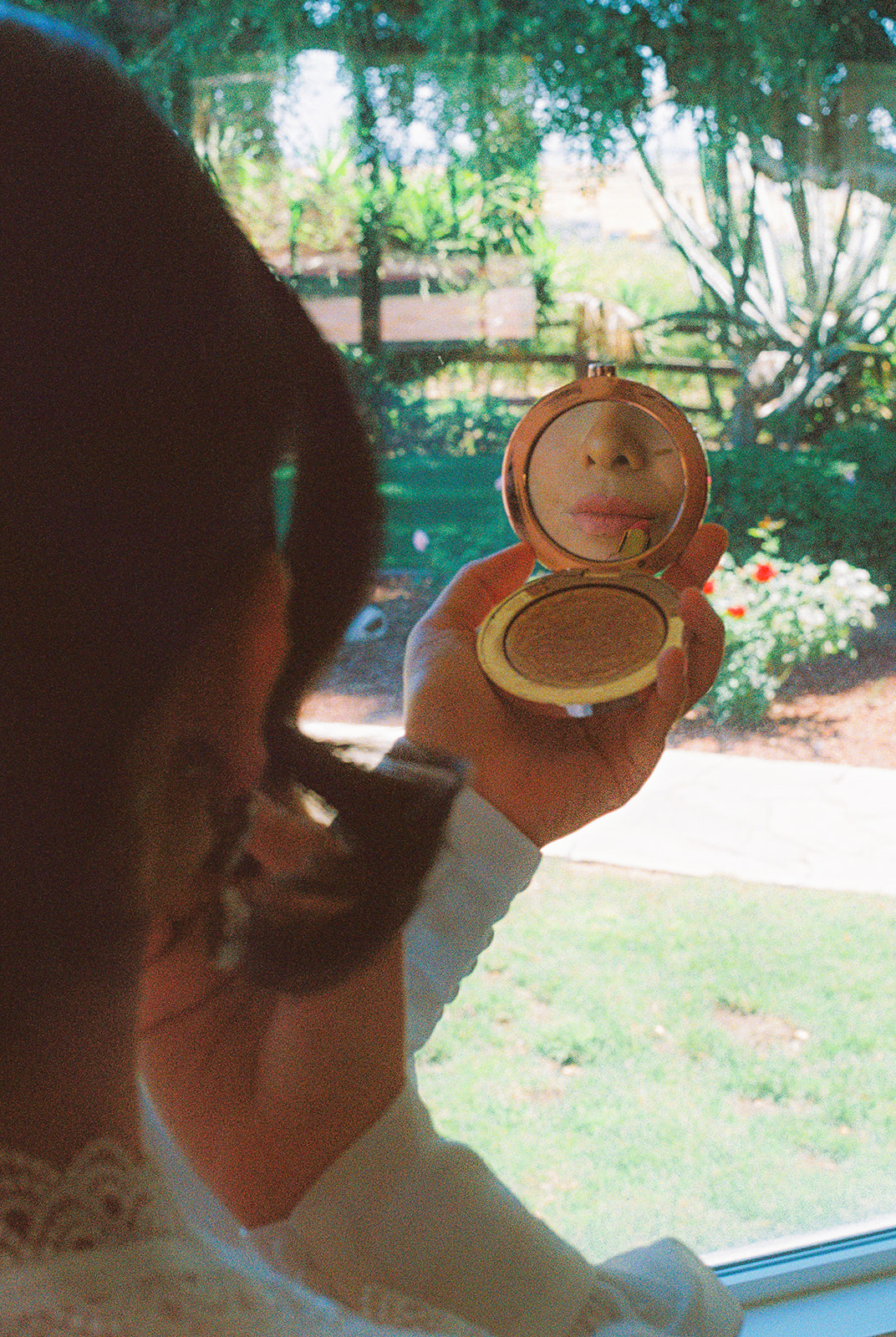 Film photograph of a bride applying lipstick in a mirror beside a window at Olive Hill Estate in Byron, California.