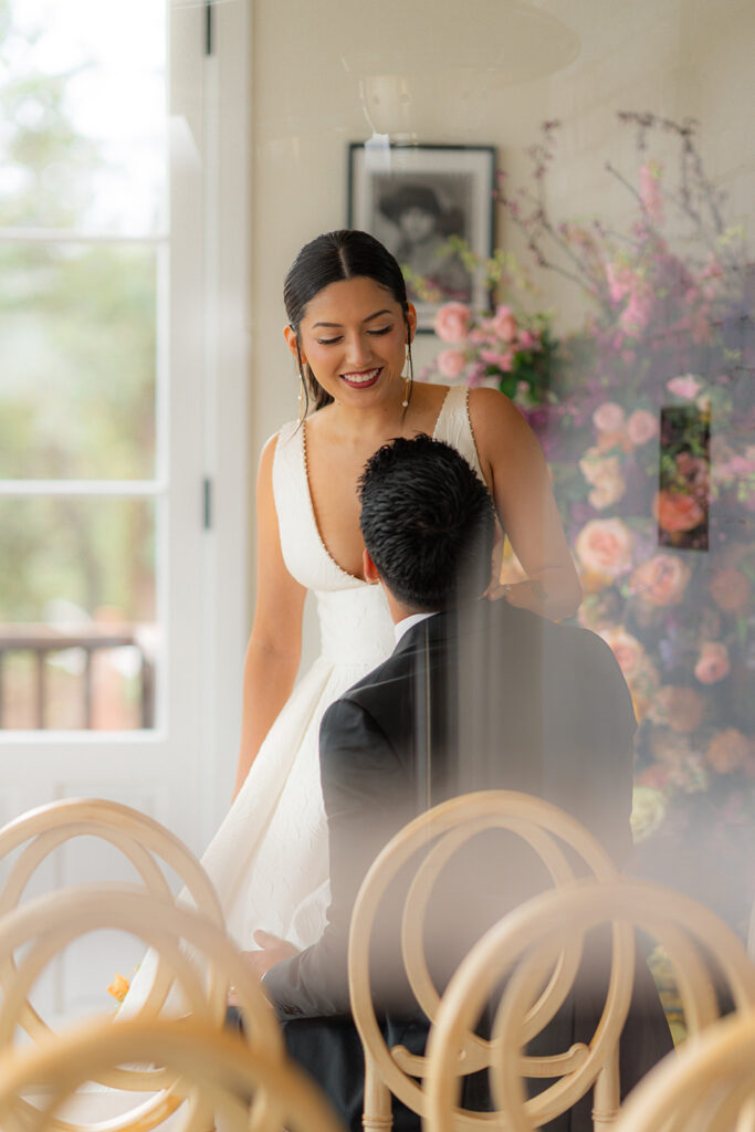 Bride and groom photographed through a window with chairs in the foreground at Hollins House in Santa Cruz, California.