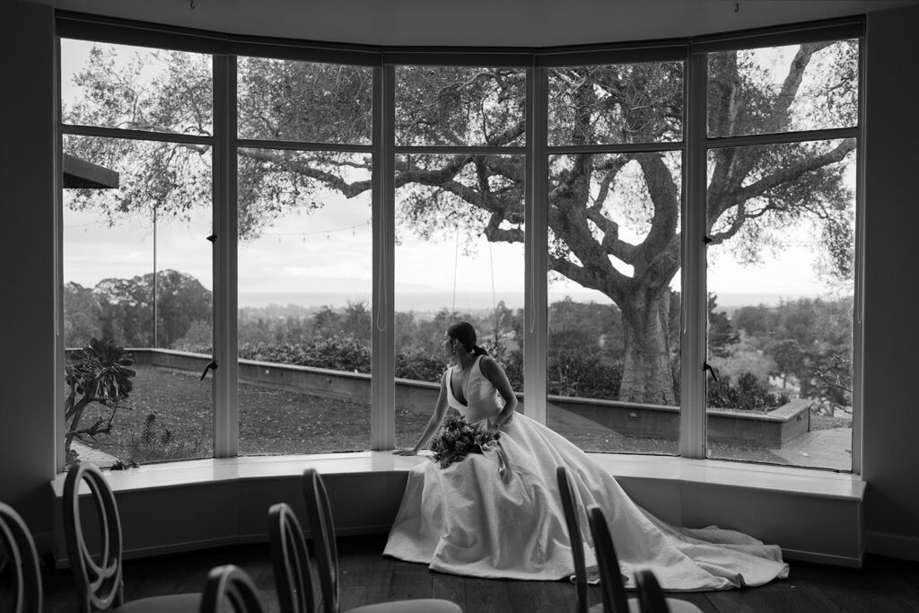 Bride sitting in a large window with a tree visible outside at Hollins House in Santa Cruz, California.