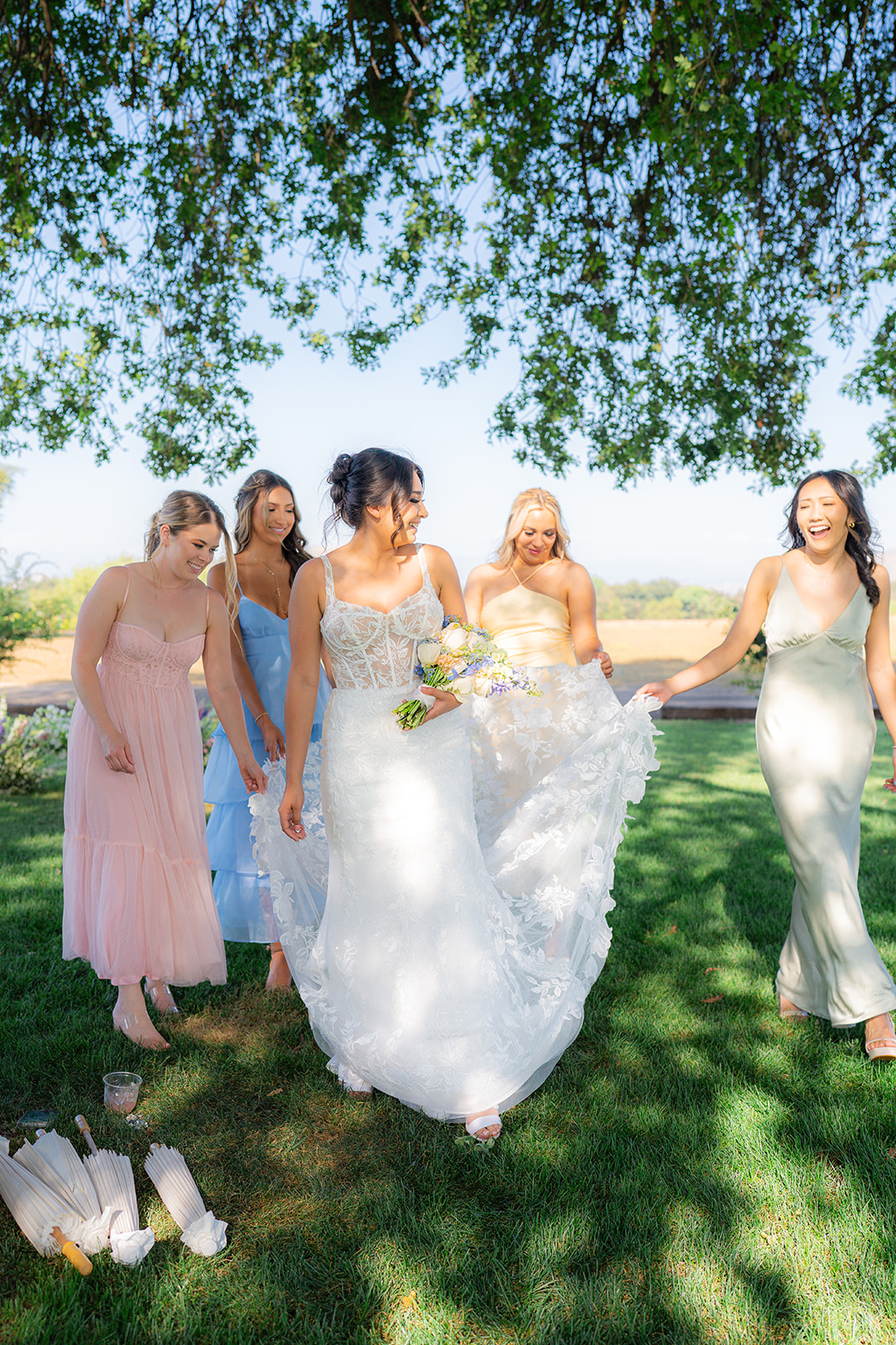 Candid moment of the bride with her bridesmaids in pastel dresses beneath a willow tree at Rainbow Oaks Ranch in Sanger, California.