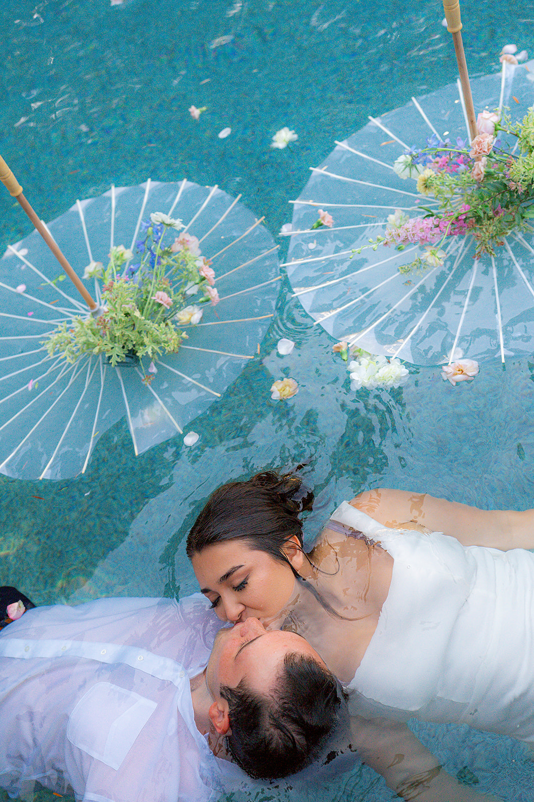 Bride and groom kissing in a pool surrounded by parasol umbrellas and flowers at Rainbow Oaks Ranch in Sanger, California.
